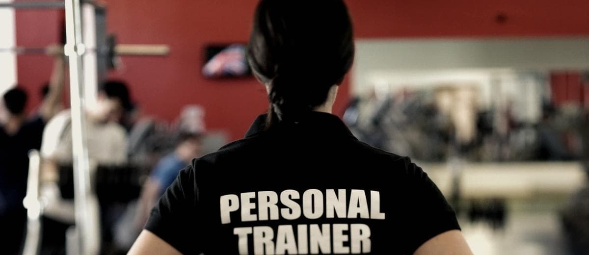 Entrenadora de espaldas con la palabra Personal Trainer en la camiseta, en el piso de un gimnasio con socios entrenando.