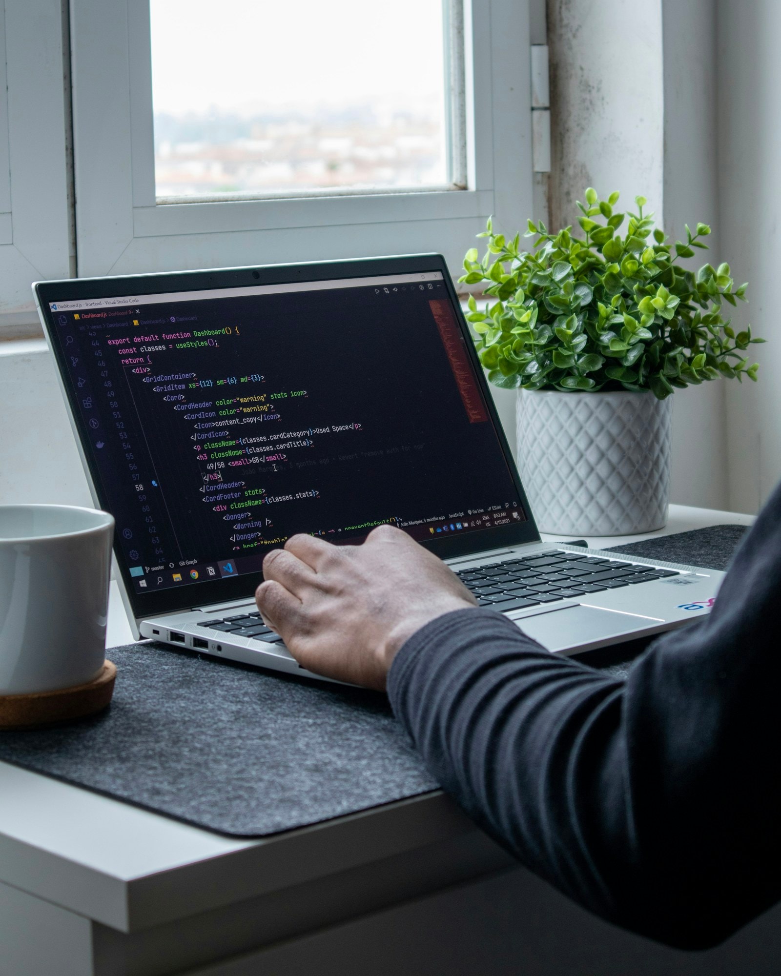 Manos sobre el teclado de un computador portátil con código en la pantalla, junto a una ventana y una planta.